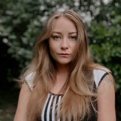 A woman with long, straight blonde hair and a black and white striped top stands against a backdrop of lush green foliage in Atlanta, Georgia.