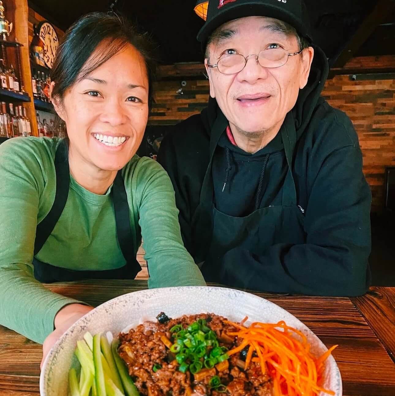 A smiling woman and man proudly present a large bowl of colorful noodles topped with ground meat, vegetables, and garnishes on a rustic wooden table in Atlanta, Georgia.