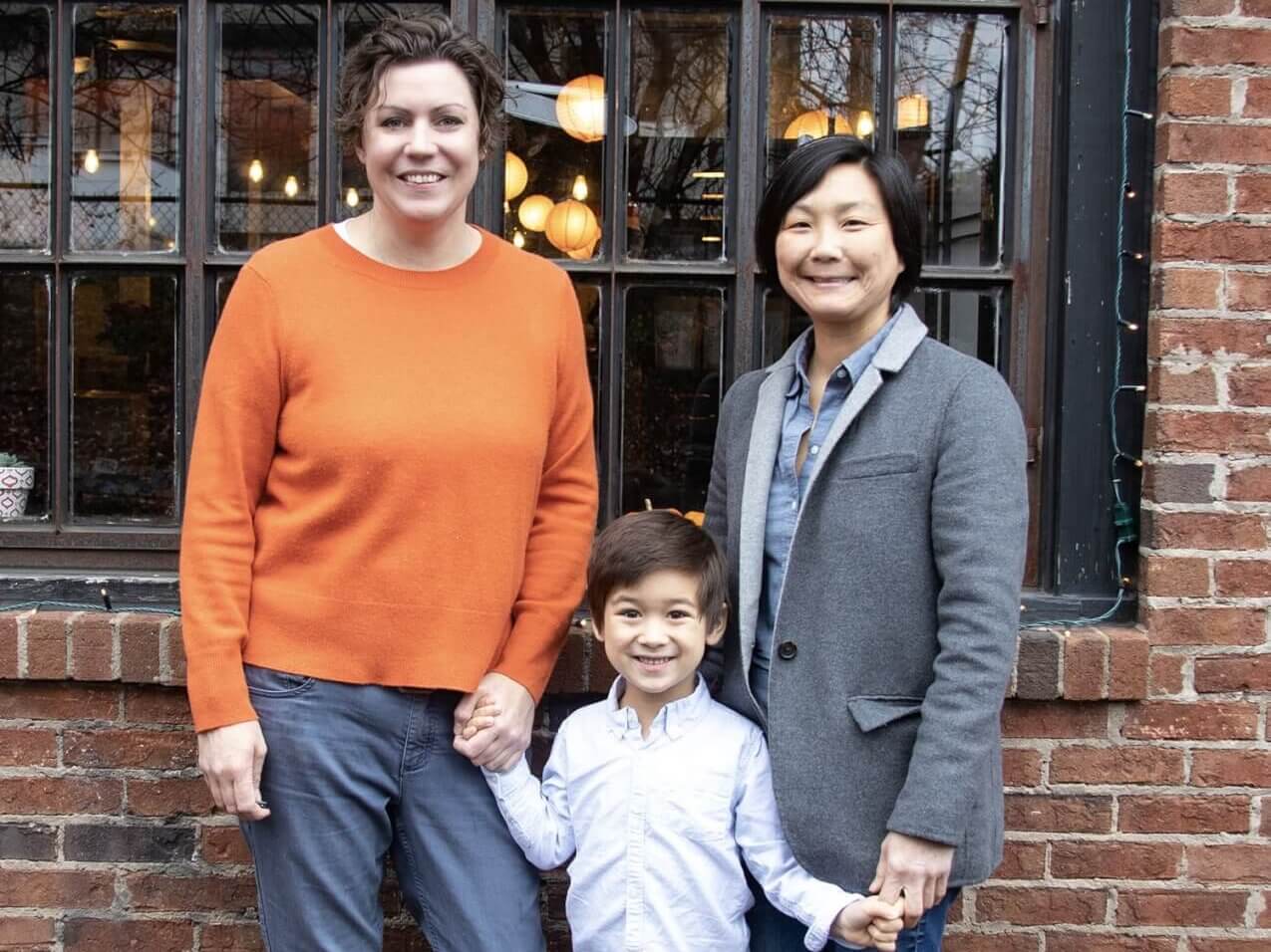 A family of three, consisting of two women and a young boy, stand hand in hand in front of a brick building with large windows and string lights in Atlanta, Georgia.