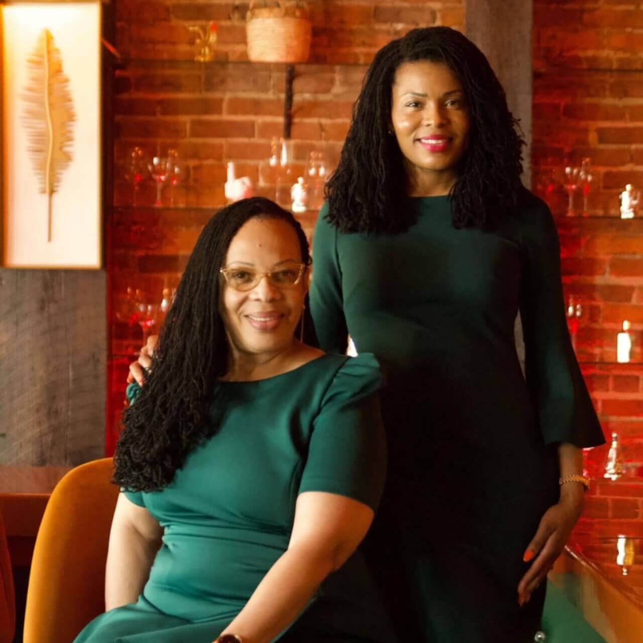 Two women in elegant green dresses smile warmly at the camera, seated and standing in a cozy, dimly lit room with a rustic brick wall and a bar in the background in Atlanta, Georgia.