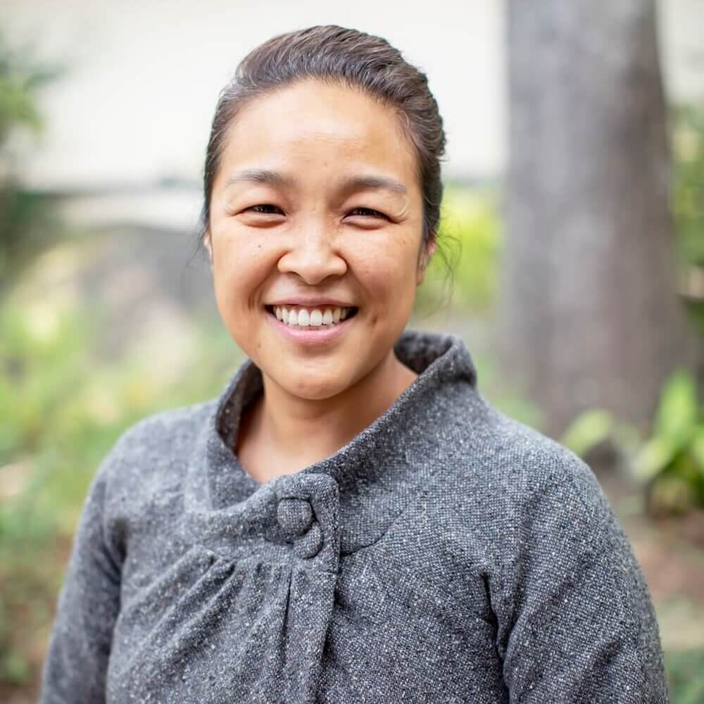 A woman with a warm smile stands outdoors, wearing a stylish gray sweater with a unique collar design in Atlanta, Georgia.