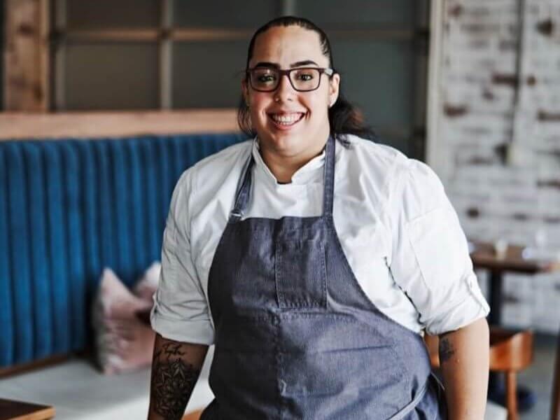 A smiling chef wearing a white coat and apron stands confidently in a restaurant kitchen with a blue booth and wooden tables in the background in Atlanta, Georgia.