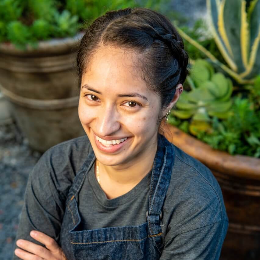 A woman with a braided hairstyle and a denim apron smiles confidently with her arms crossed in Atlanta, Georgia.