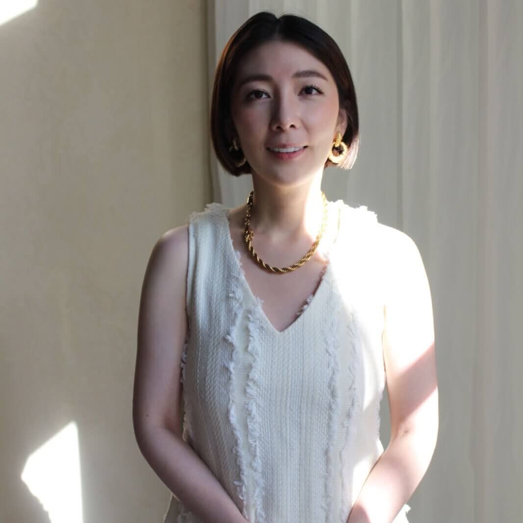 A woman in a white dress stands with her hands clasped in front of her, wearing gold jewelry against a light-colored background in Atlanta, Georgia.