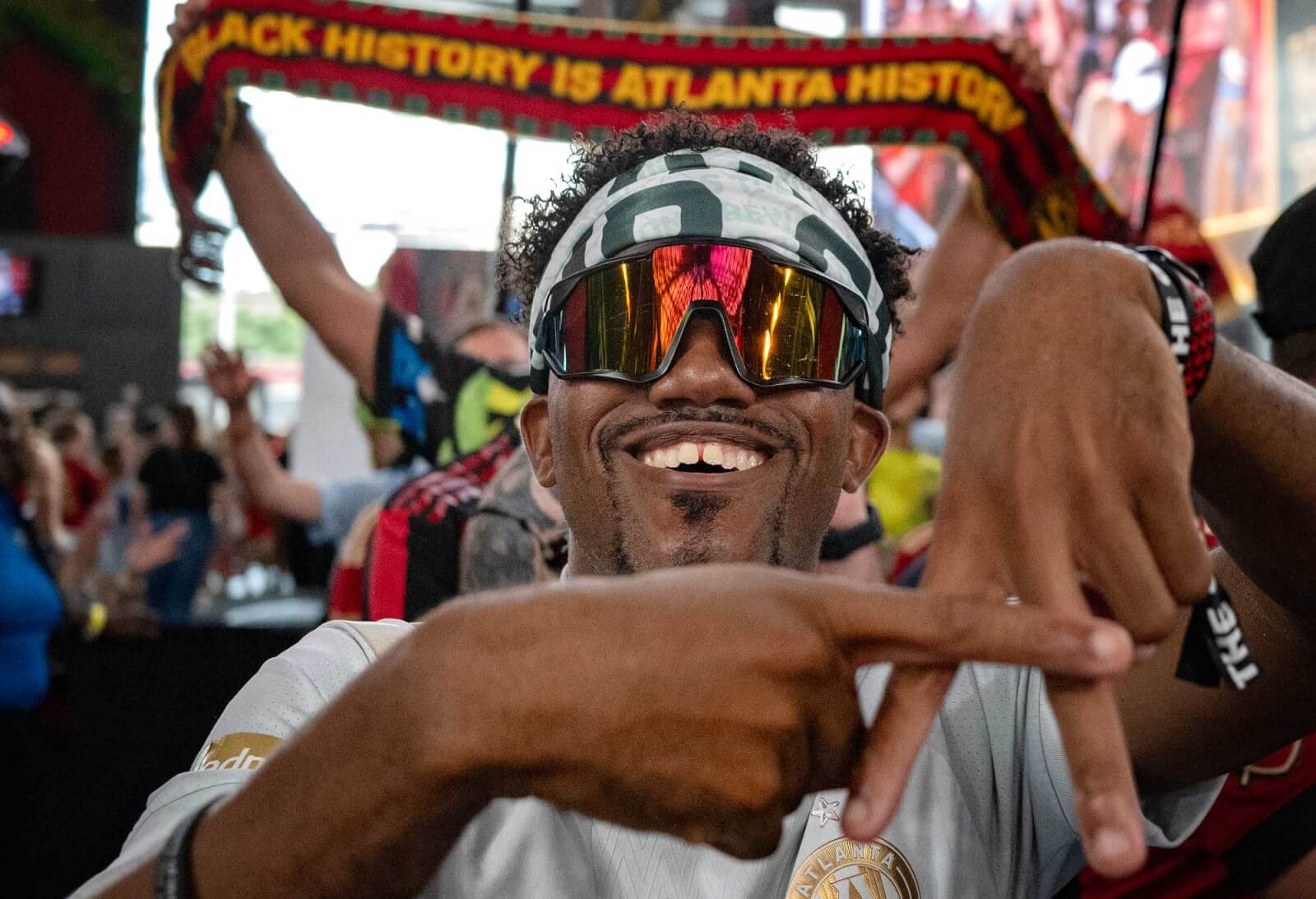 A man wearing a headband and sunglasses with reflective lenses smiles broadly and makes a peace sign with both hands in Atlanta, Georgia.