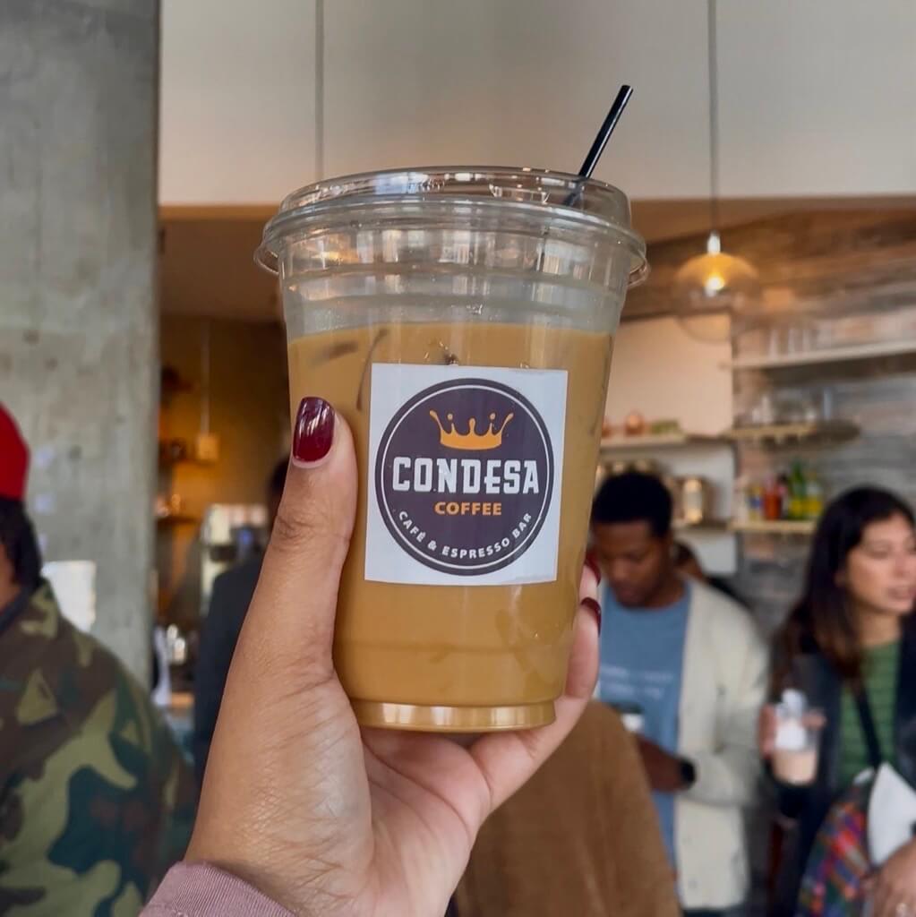 One Day Neighborhood Hopping Around Atlanta 1 A hand holds a clear plastic cup filled with iced coffee from Condesa Coffee, featuring a crown logo and a black straw in Atlanta, Georgia.