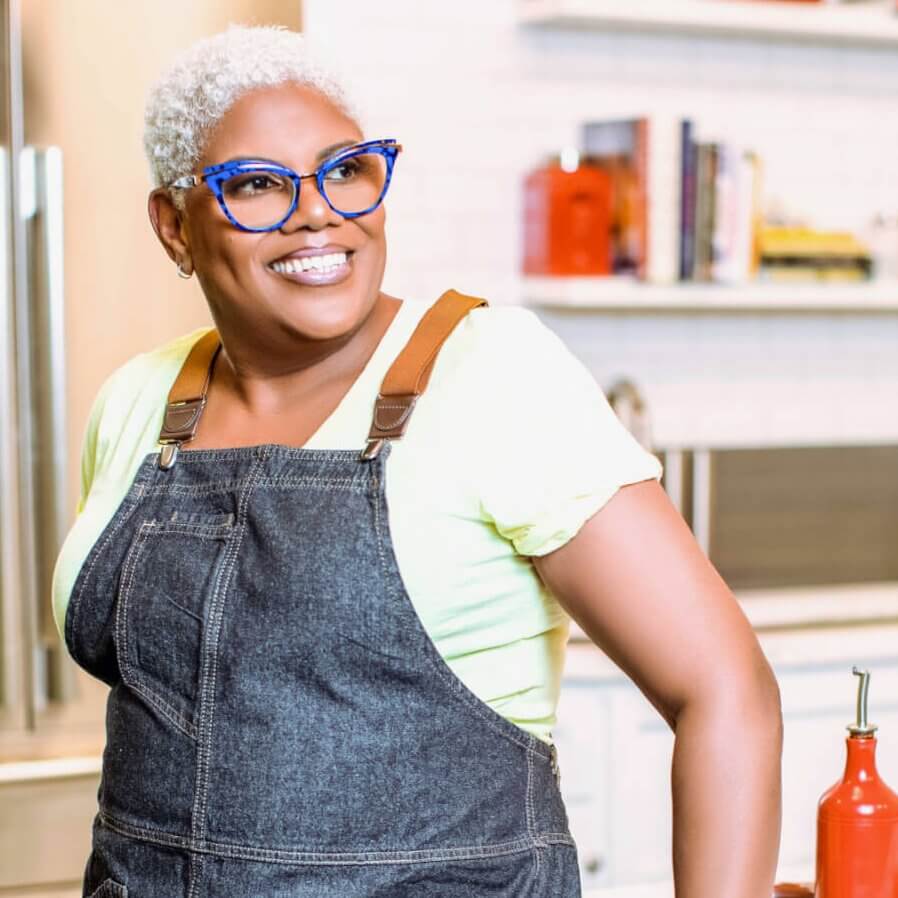 A woman with short white hair, wearing blue-framed glasses, a yellow t-shirt, and denim overalls, stands smiling in a modern kitchen with white cabinets and red accents in Atlanta, Georgia.