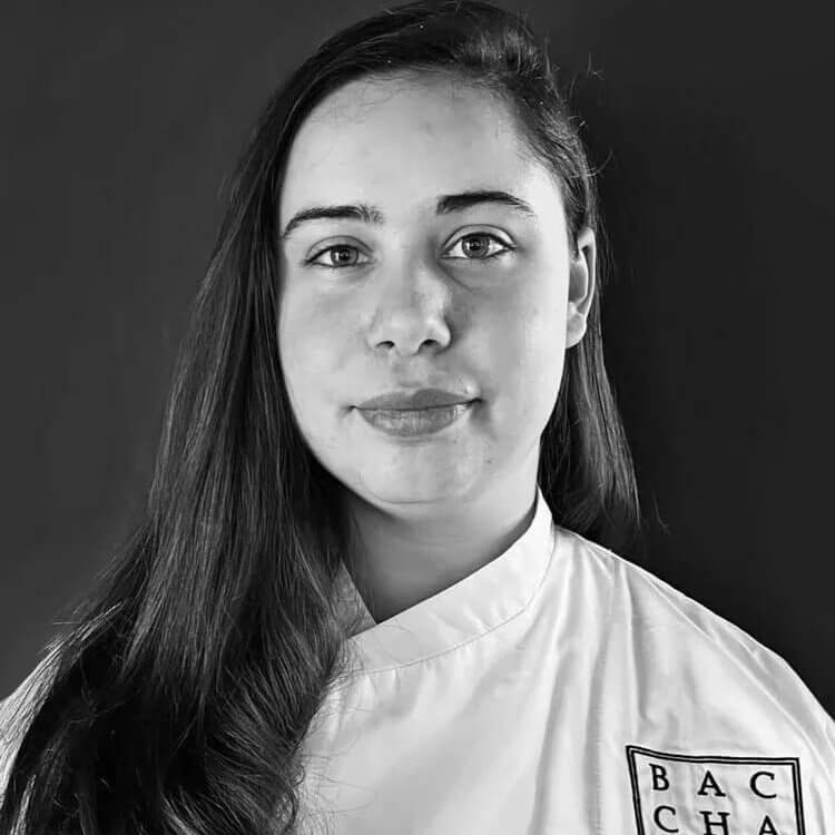 A young woman with long, dark hair smiles gently, wearing a white chef's jacket with the logo 'BACCHANALIA' on the sleeve in Atlanta, Georgia.