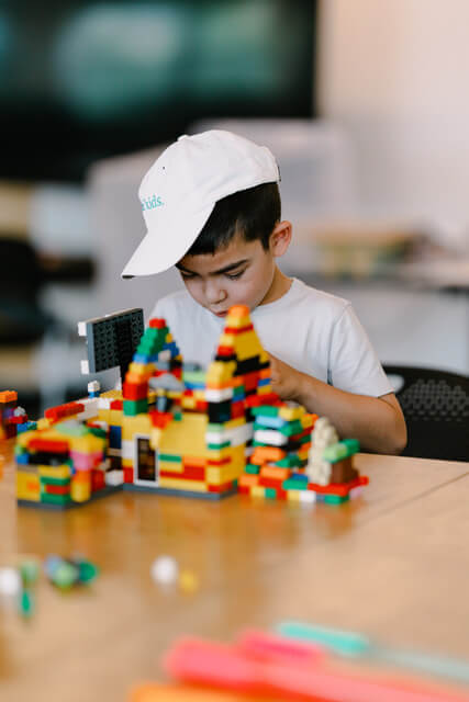 A young boy wearing a white cap is deeply focused on building a colorful LEGO structure on a wooden table in Atlanta, Georgia.