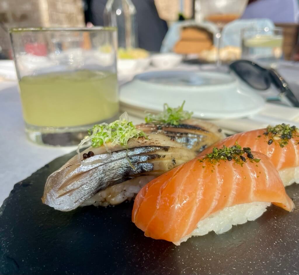 Three pieces of sushi, including two salmon nigiri and one grilled fish nigiri, are artfully arranged on a black slate plate, accompanied by a glass of green juice and a bowl of soup in Atlanta, Georgia.