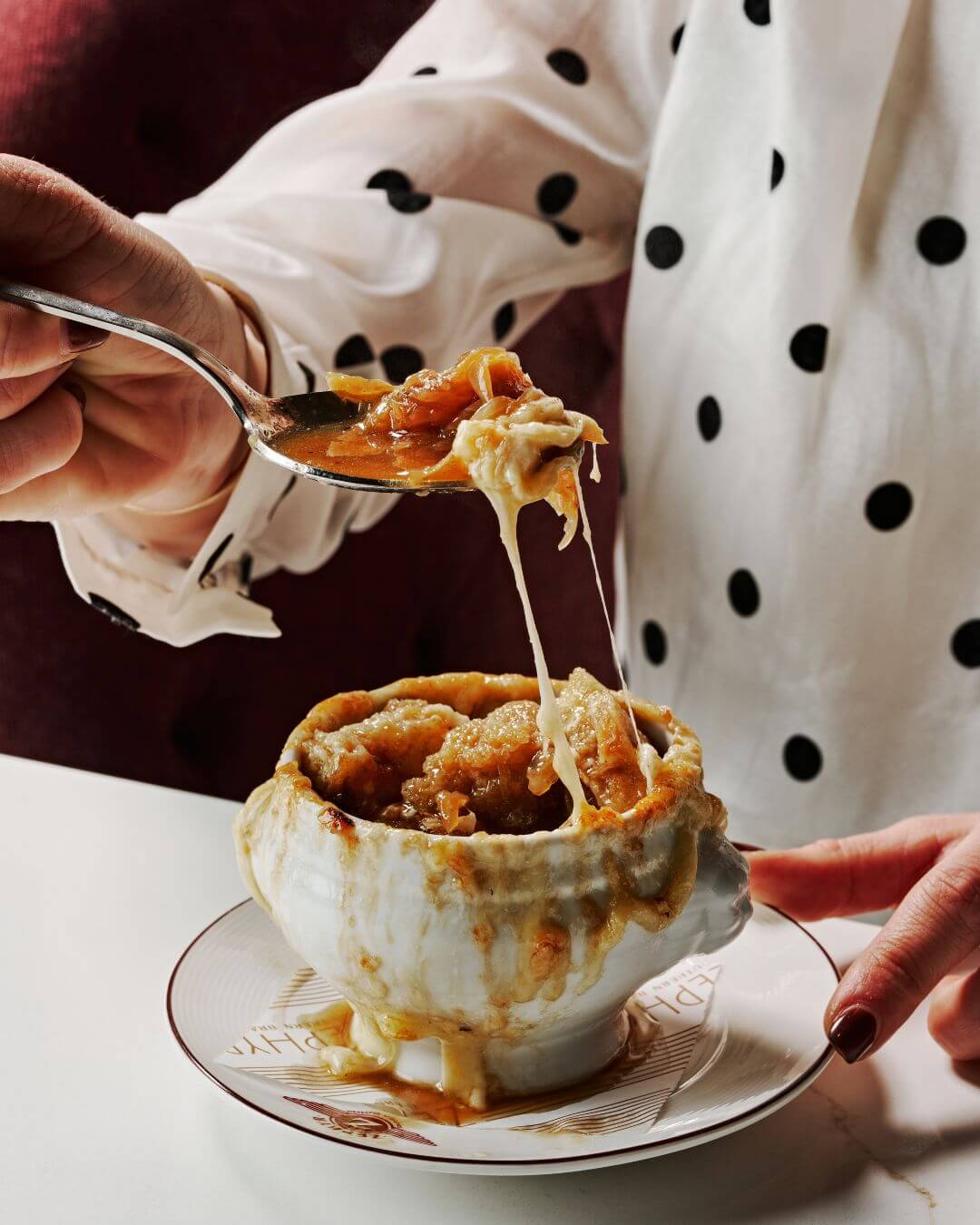 Ultimate Guide to Hotel Dining ATL Style 8 A person in a polka-dotted blouse is holding a spoonful of melted cheese from a bowl of French onion soup, which is served in a white bowl with a golden rim on a matching saucer in Atlanta, Georgia.