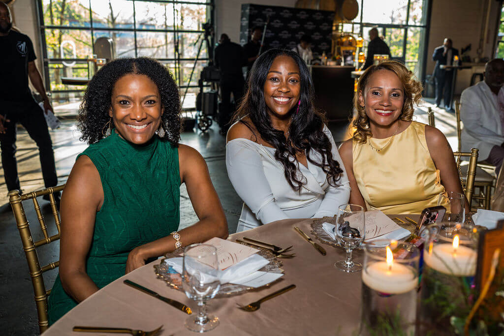 Three elegantly dressed women smile warmly at the camera while seated at a beautifully set table with golden flatware and lit candles in Atlanta, Georgia.