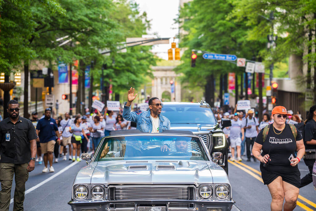A man waves from a classic convertible car as a large crowd marches down a city street lined with trees and buildings in Atlanta, Georgia.