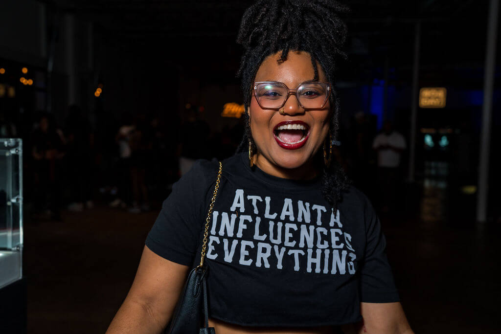 A woman with dreadlocks and glasses wearing a black t-shirt that reads 'Atlanta Influences Everything' smiles broadly in Atlanta, Georgia.