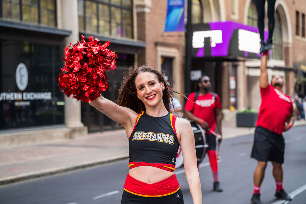 A cheerleader in a black and red Skyhawks uniform waves a red pom-pom in the air during a parade in Atlanta, Georgia.