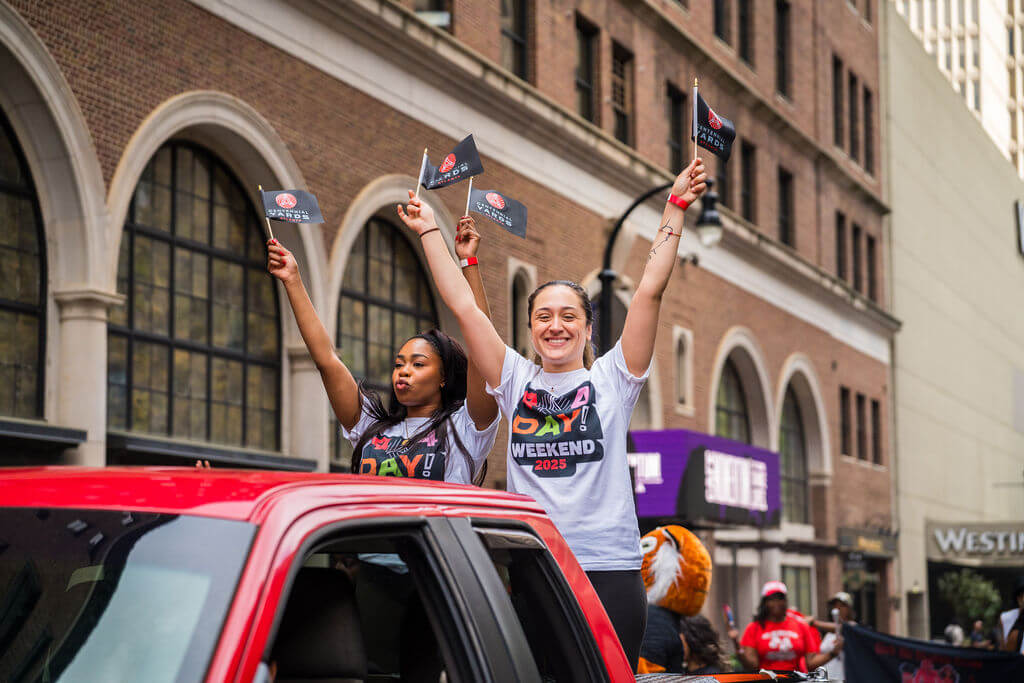 Two women joyfully wave flags from the back of a red pickup truck during a parade, surrounded by cheering crowds and colorful buildings in Atlanta, Georgia.
