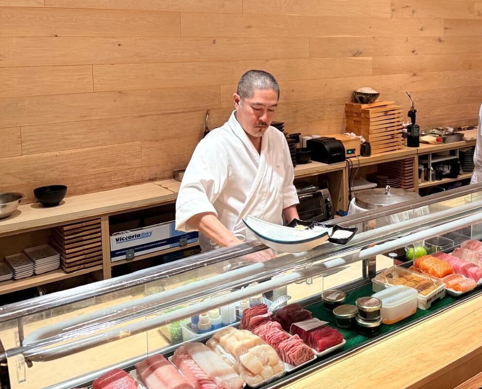 A skilled sushi chef in a white kimono prepares fresh sashimi behind a glass display case in a modern Japanese restaurant in Atlanta, Georgia.