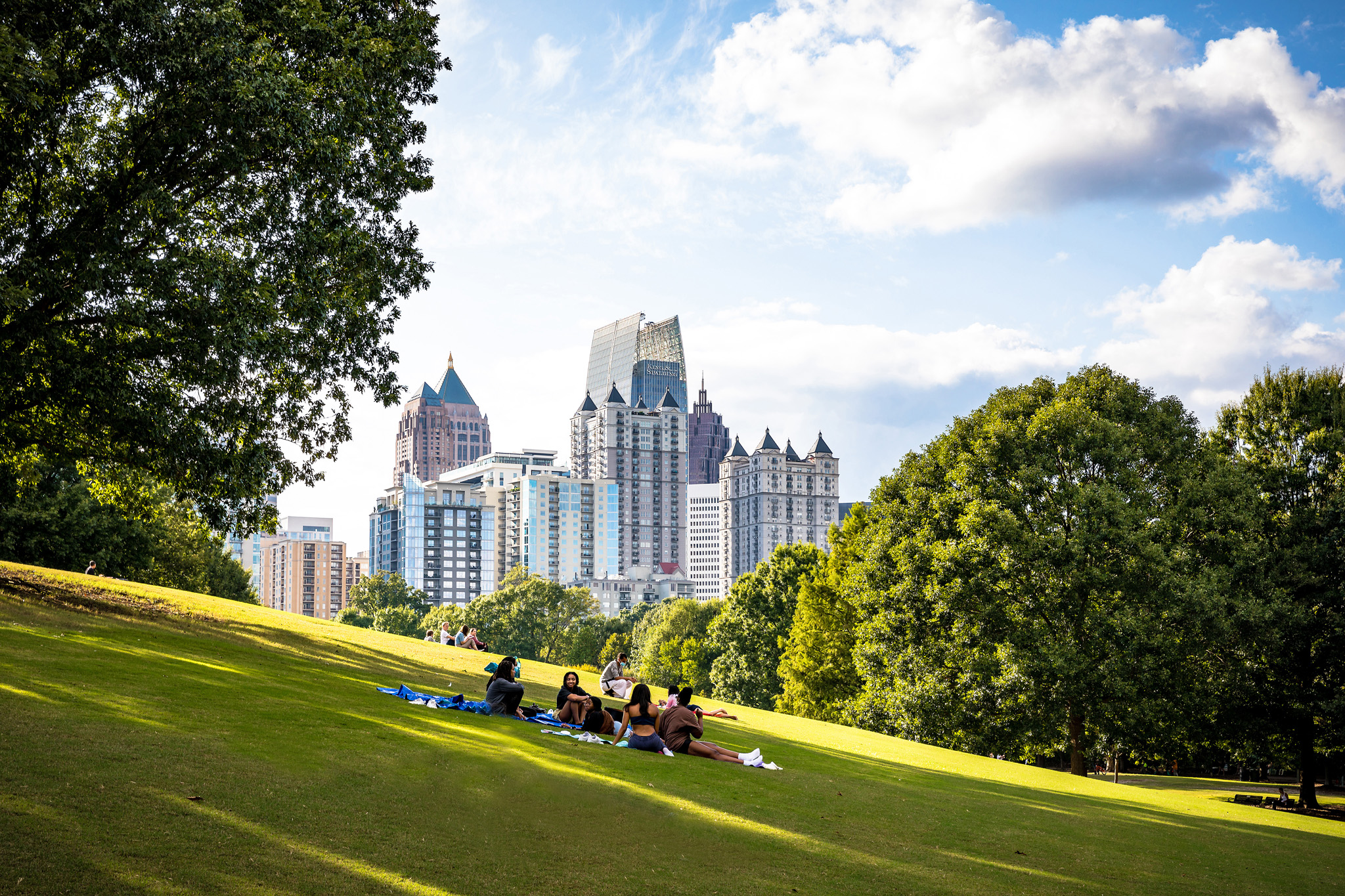 People relax on a grassy hill in a park, with a city skyline visible in the distance in Atlanta, Georgia.
