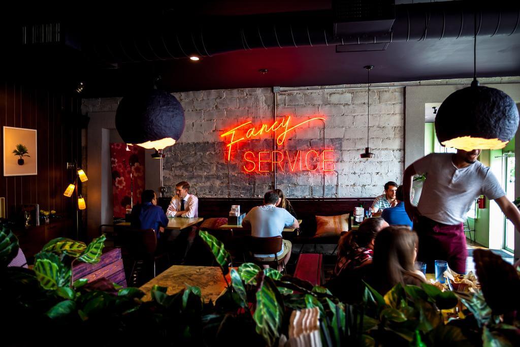 A dimly lit restaurant with a neon sign reading 'Fancy Service' and patrons seated at tables in Atlanta, Georgia.