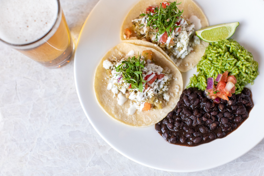 A plate of two tacos topped with fresh greens, cheese, and salsa, served with black beans, rice, and a lime wedge, accompanied by a glass of beer in Atlanta, Georgia.