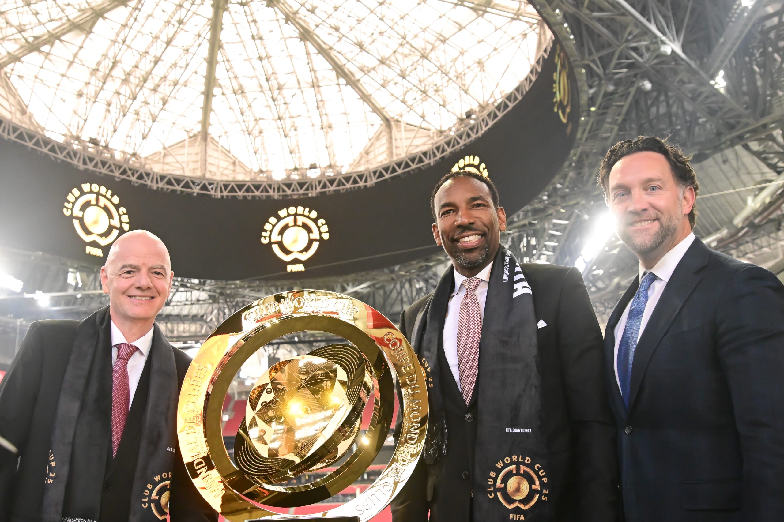Three men in suits pose with a large golden trophy in a domed stadium in Atlanta, Georgia.