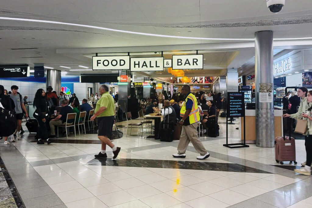 Travelers and airport staff move through a bustling airport terminal, passing by a prominent 'Food Hall & Bar' sign in Atlanta, Georgia.