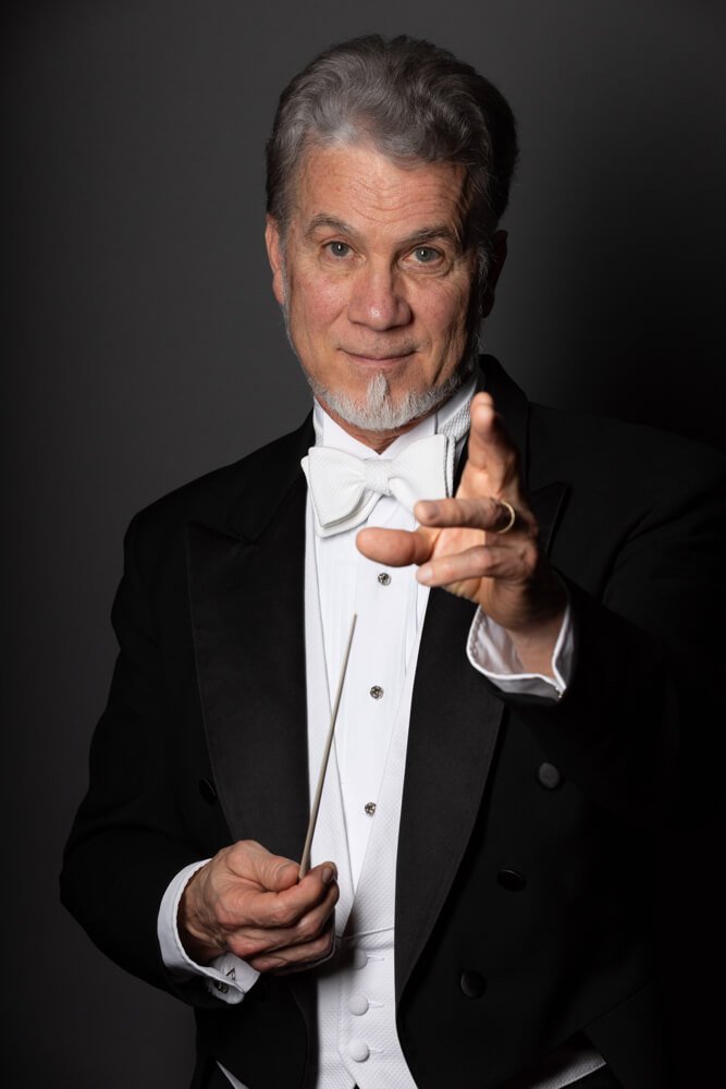 "Frida" Comes to the Stage at the Atlanta Ballet 6 A distinguished conductor dressed in a formal black tuxedo with a white bow tie and shirt, holding a baton in his right hand, gestures with his left hand against a dark background in Atlanta, Georgia.