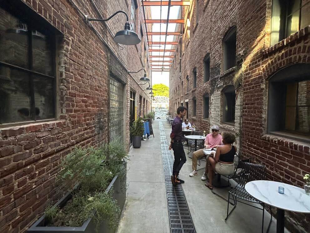 A narrow alleyway between brick buildings features a covered outdoor seating area with tables and chairs, where people are enjoying their time in Atlanta, Georgia.