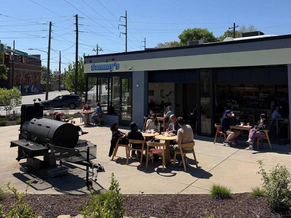 A group of people are enjoying a sunny day at Sammy's, a casual outdoor dining area with picnic tables, a large grill, and a clear blue sky overhead in Atlanta, Georgia.