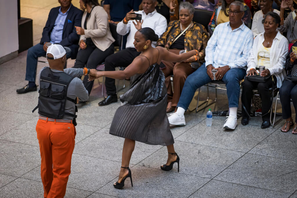 A woman in a black dress and high heels dances with a man in orange pants and a vest in front of an audience seated on chairs in Atlanta, Georgia.