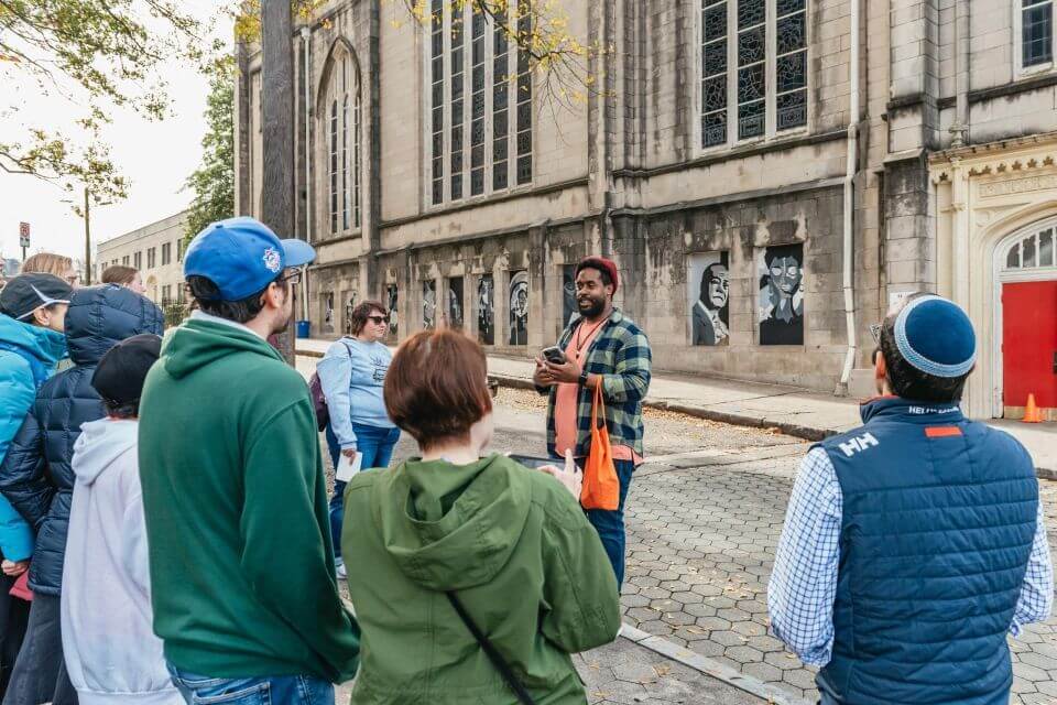 A diverse group of people, some wearing winter jackets and hats, are gathered on a sidewalk in front of a historic building with large windows and murals, listening to a tour guide speaking into a microphone in Atlanta, Georgia.