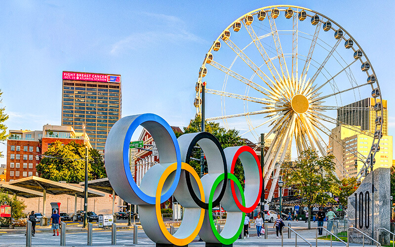 The Olympic rings stand tall in front of a vibrant cityscape featuring a towering Ferris wheel and modern skyscrapers in Atlanta, Georgia.