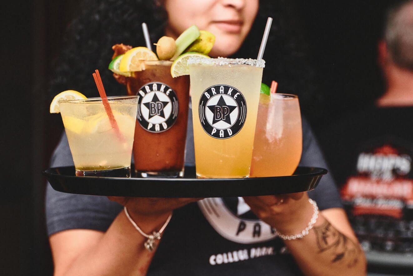 A bartender holds a tray with five colorful cocktails, each garnished with fruit and branded with the 'Brake Pad' logo in Atlanta, Georgia.