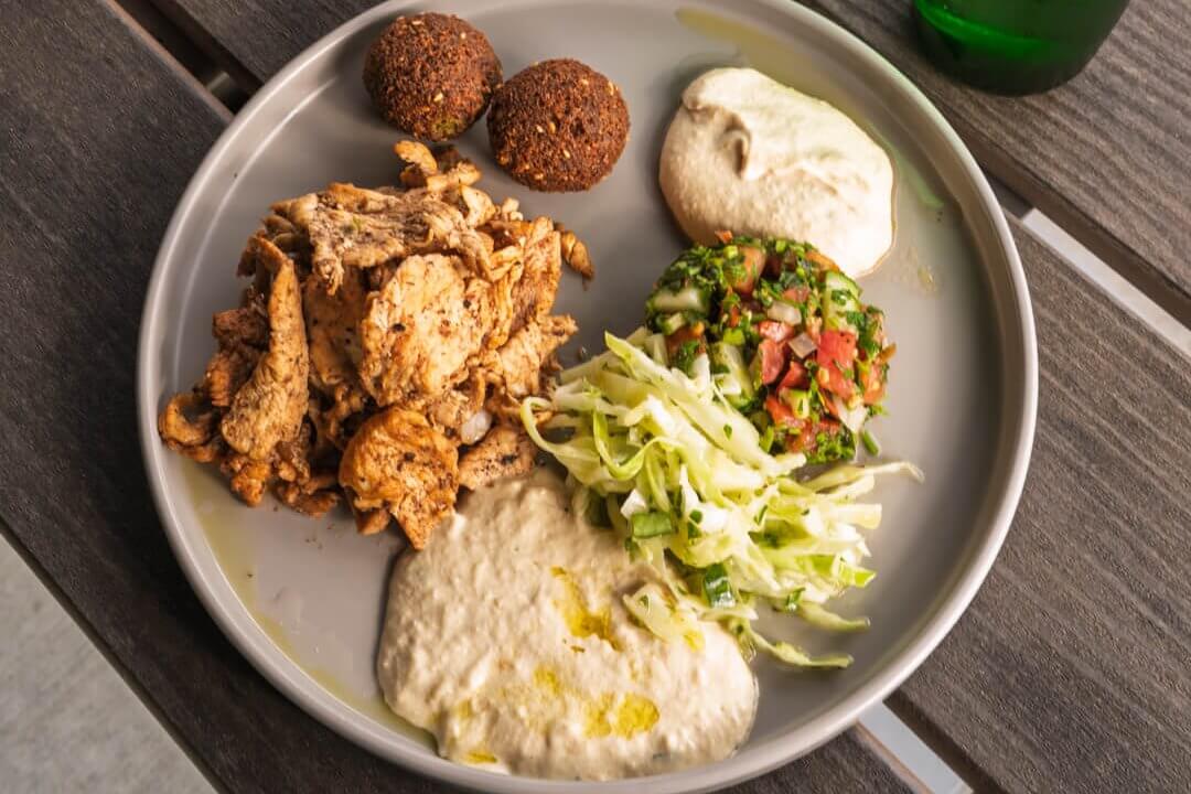 A colorful Mediterranean-style meal featuring grilled chicken, falafel balls, hummus, tabbouleh salad, and baklava dessert, accompanied by a bottle of Perrier sparkling water in Atlanta, Georgia.