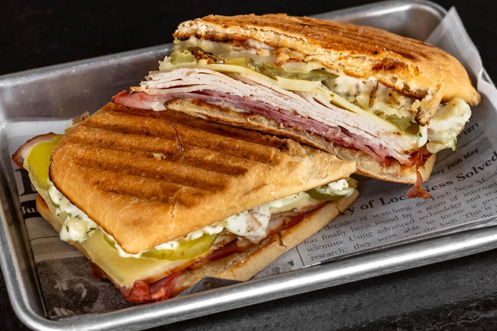 A mouthwatering Cuban sandwich with toasted bread, layers of ham, turkey, cheese, pickles, and mustard, served on a metal tray lined with newspaper in Atlanta, Georgia.