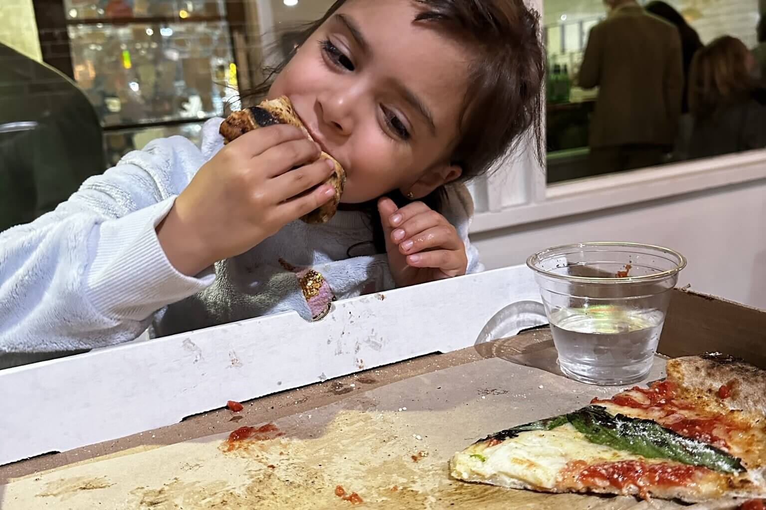 A young girl enjoys a slice of pizza with toppings, sitting at a table with a glass of water in Atlanta, Georgia.