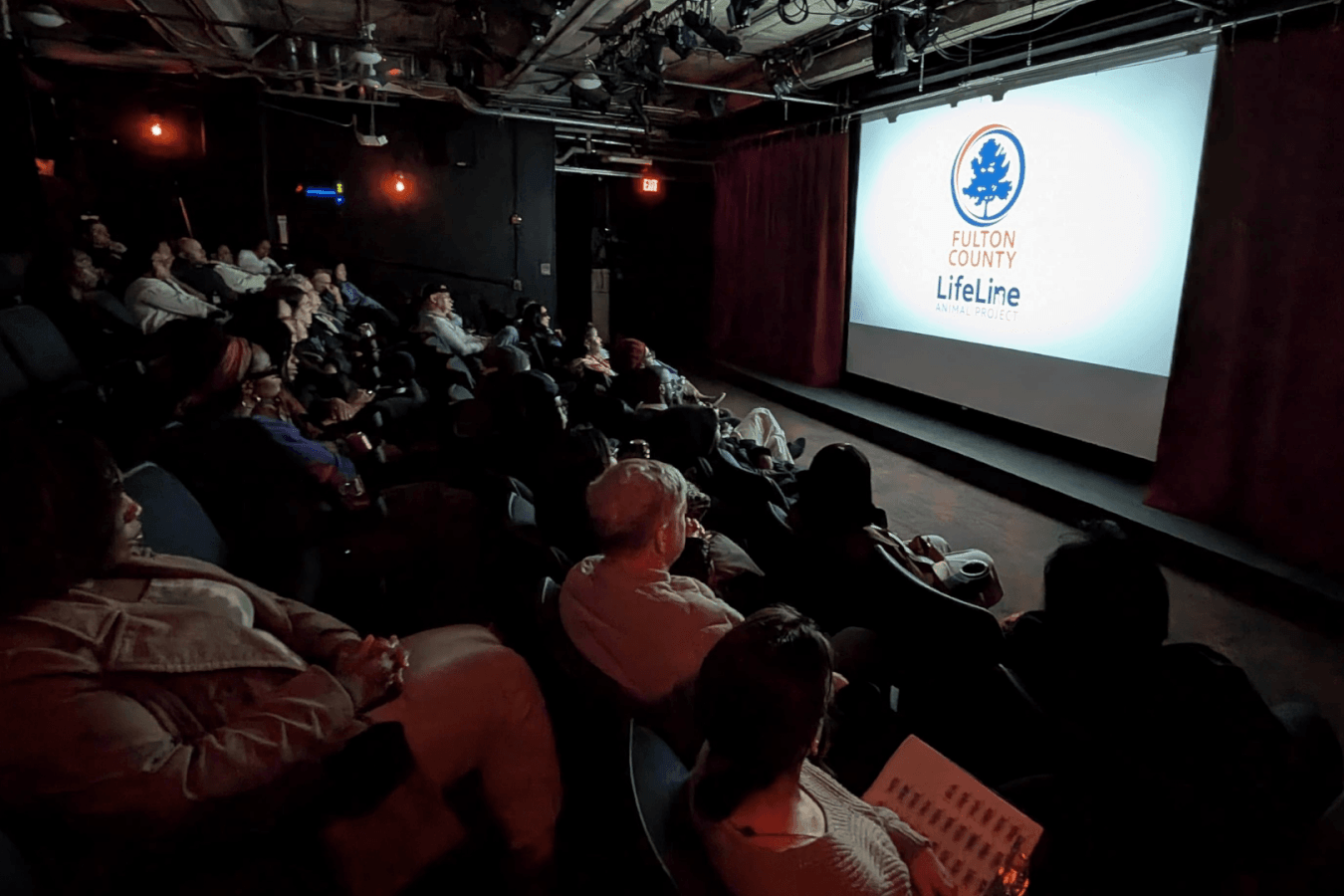 An audience is seated in a dimly lit theater, watching a presentation on a large screen that displays the Fulton County Lifeline logo in Atlanta, Georgia.