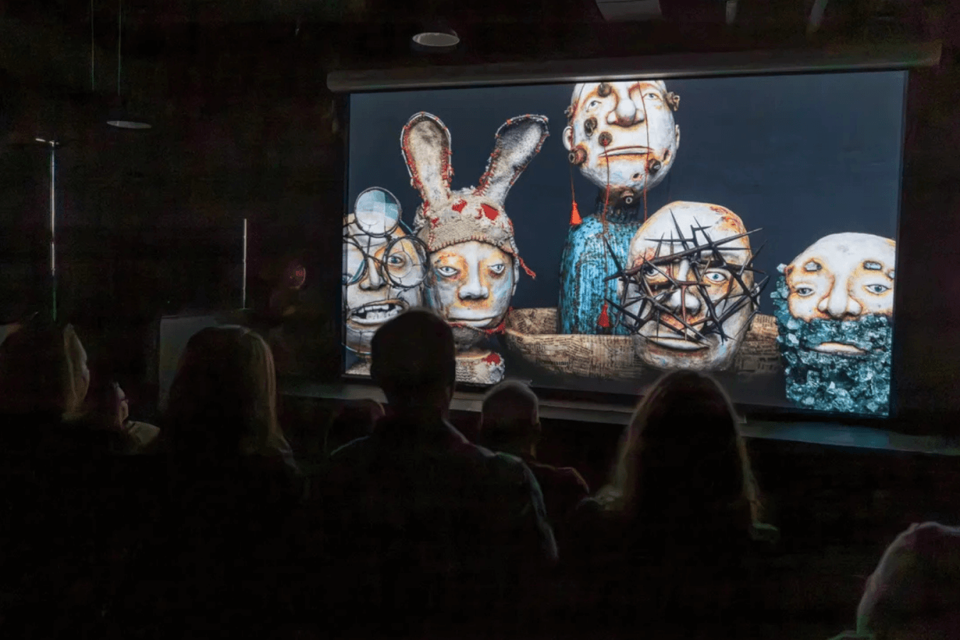 An audience sits in a dark theater, watching a large screen displaying a collection of surreal, mask-like faces with exaggerated features and intricate designs in Atlanta, Georgia.