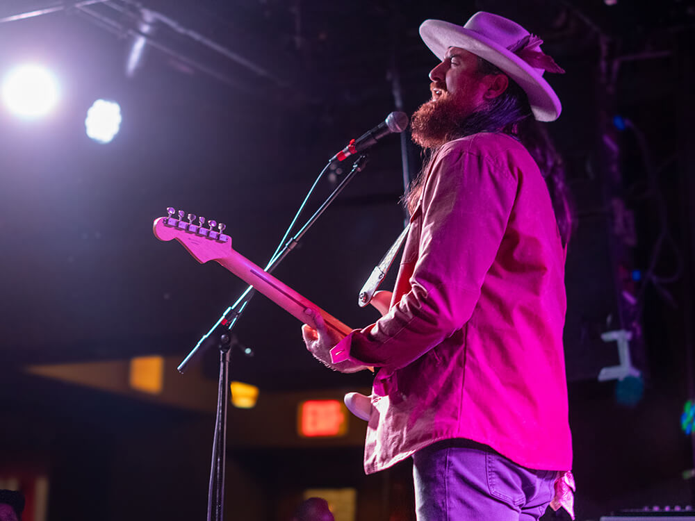 A musician wearing a wide-brimmed hat and a pink jacket performs on stage with an electric guitar, illuminated by stage lights in Atlanta, Georgia.