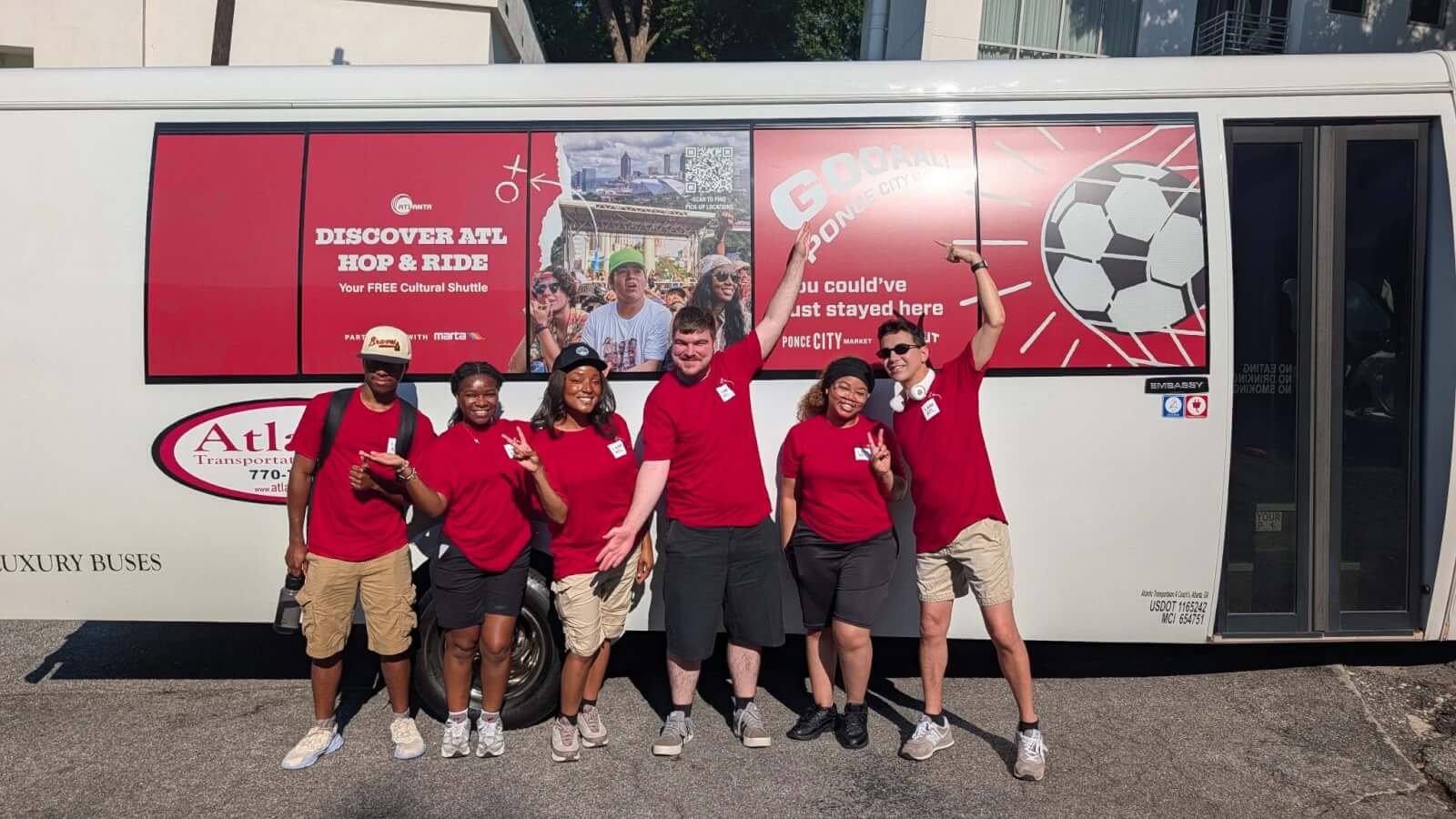 A group of six young people, all wearing red shirts, pose in front of a red and white bus with the text 'Discover ATL Hop & Ride' and a soccer ball graphic in Atlanta, Georgia.