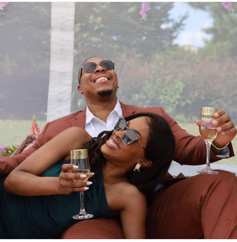 A couple in matching brown suits enjoys champagne on a grassy area with floral decorations in Atlanta, Georgia.