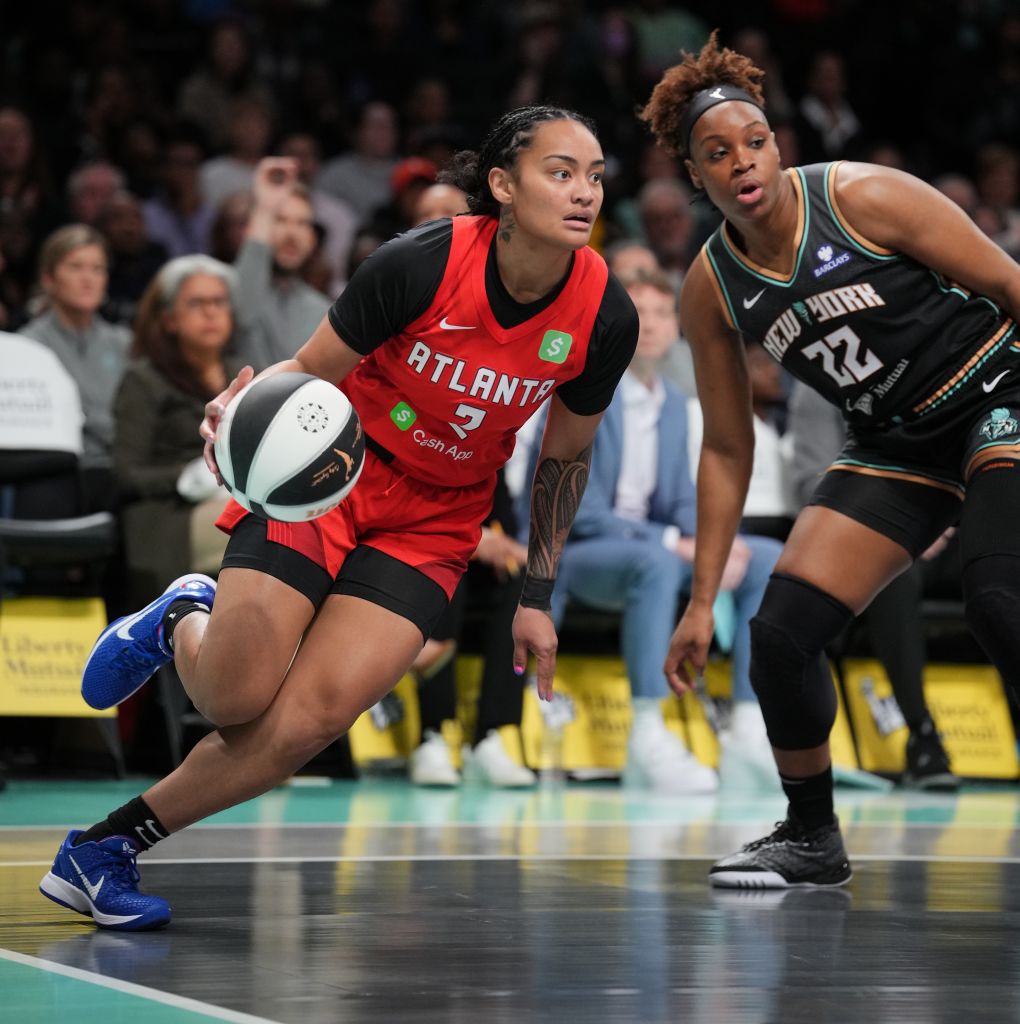 Two women basketball players are in action on the court, with one player in a red Atlanta jersey dribbling the ball while being closely guarded by an opponent in a black New York jersey in Atlanta, Georgia.