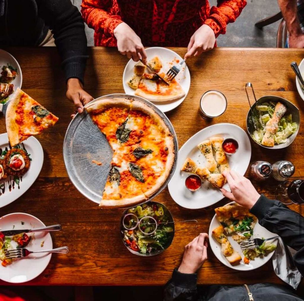 A group of people are gathered around a wooden table, enjoying a variety of Italian dishes including pizza, pasta, salads, and breadsticks, with drinks in hand in Atlanta, Georgia.