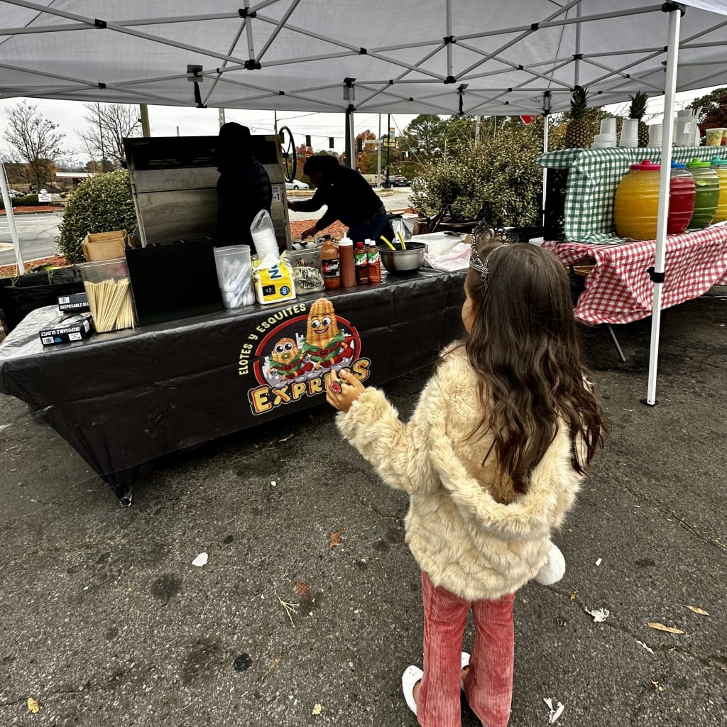 A young girl in a fluffy coat stands near a food truck tent, watching workers prepare food in Atlanta, Georgia.