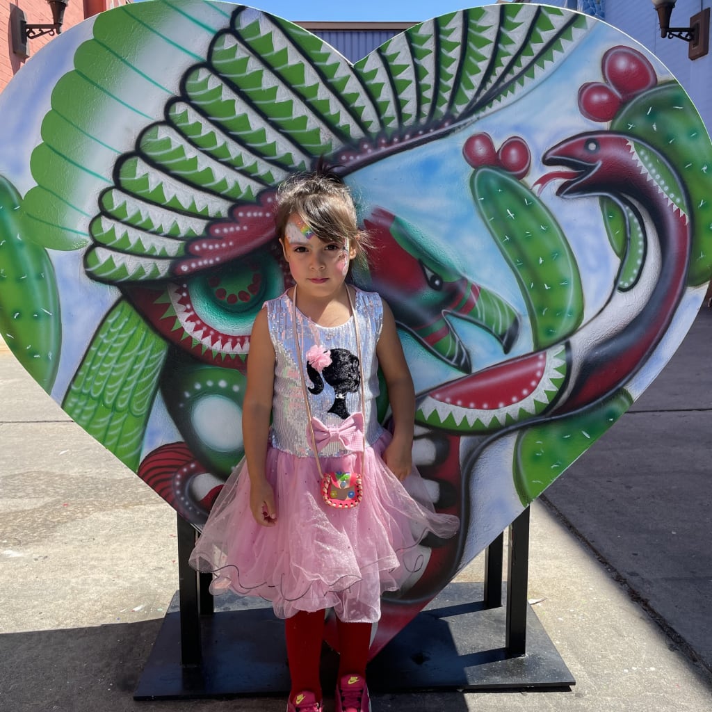 A young girl in a pink tutu stands in front of a large, colorful heart mural with Mexican folk art designs in Atlanta, Georgia.