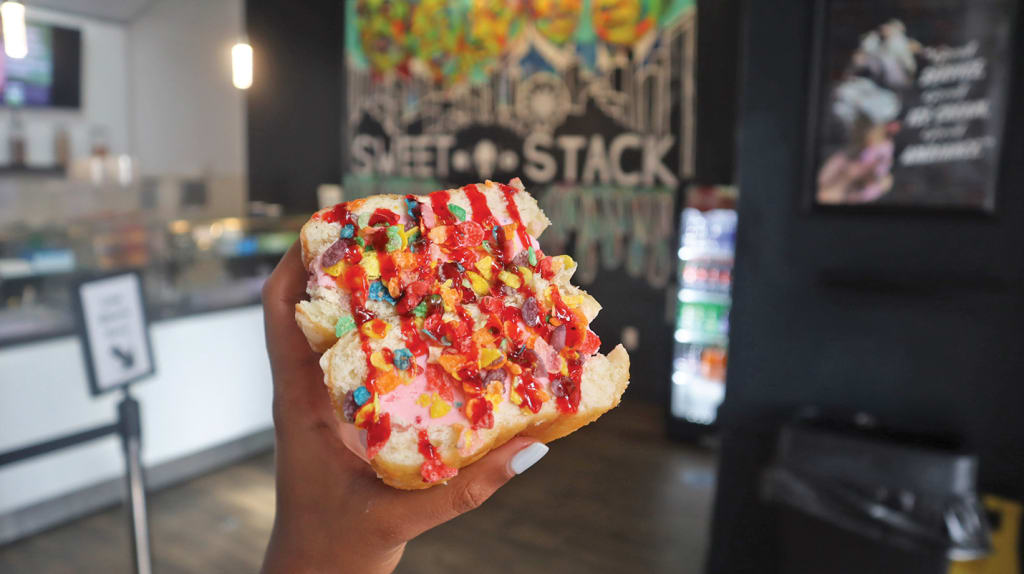 A hand holds a colorful, rainbow-sprinkled ice cream sandwich with red sauce in Atlanta, Georgia.