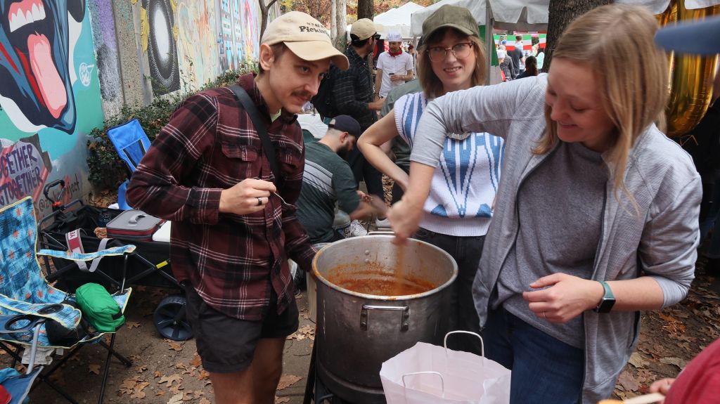 Two people stir a large pot of chili at an outdoor event with colorful graffiti in the background in Atlanta, Georgia.