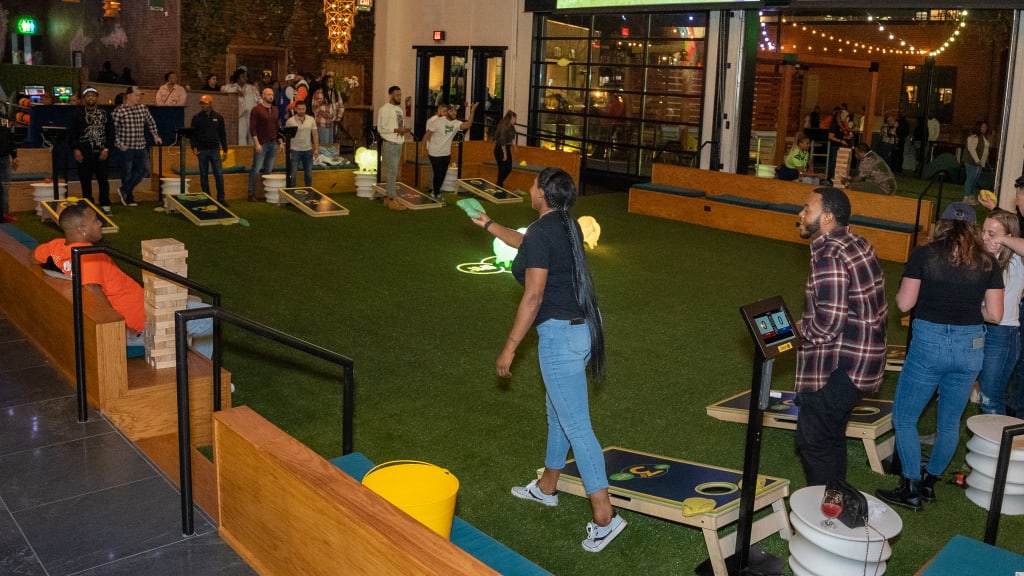 People playing cornhole and other games in a brightly lit indoor space with a green artificial turf floor in Atlanta, Georgia.