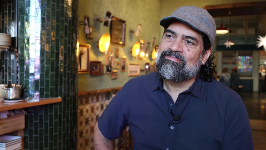 A bearded man with a gray cap stands in a cozy room with green tiles and framed pictures in Atlanta, Georgia.