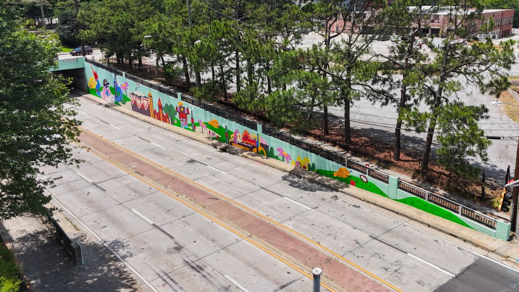 A colorful mural depicting various scenes and characters adorns a wall alongside a tree-lined road in Atlanta, Georgia.